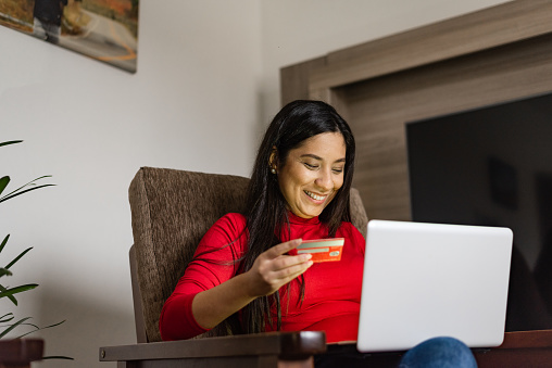 Young deaf woman sitting on the armchair holding a credit card for online shopping on laptop