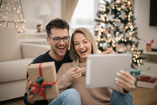 Cheerful young Caucasian couple having fun at home together, celebrating winter holidays with Christmas presents, smiling and talking to their friends and family using a digital tablet computer