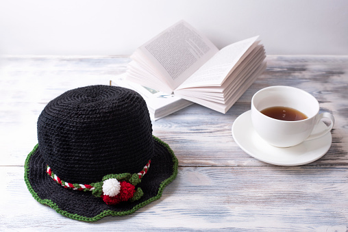 A Black knitted hat with Christmas decor is on a white wooden table with books and a cup of tea.