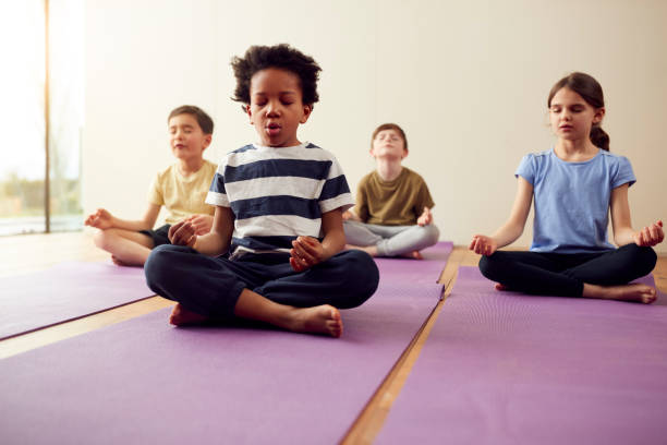 grupo de niños sentados en alfombrillas de ejercicio y meditando en el estudio de yoga - yoga fotografías e imágenes de stock