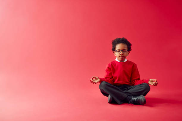赤いスタジオの背景に対して座って瞑想する制服を着た男性の小学生 - child 6 7 years education school ストックフォトと画像