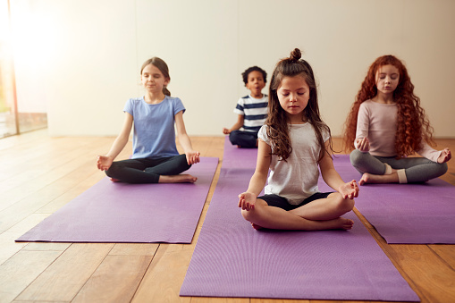 Group Of Children Sitting On Exercise Mats And Meditating In Yoga Studio