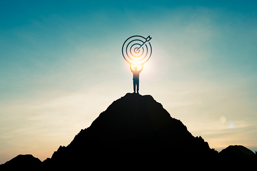 Silhouette of businessman holding target board on the top of mountain with over blue sky and sunlight. It is symbol of leadership successful achievement with goal and objective target.