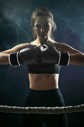 confident athletic brunette woman in boxing gloves and sportswear on dark background with smoke.
