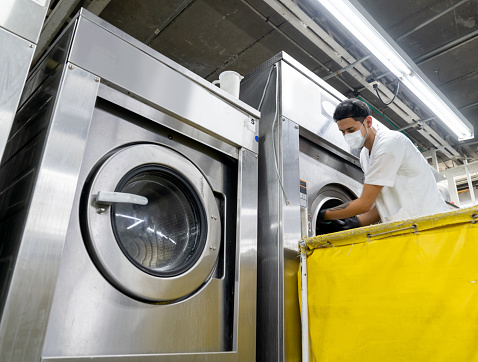 Latin American man working at the laundromat wearing a facemask and loading a washing machine during the COVID-19 pandemic