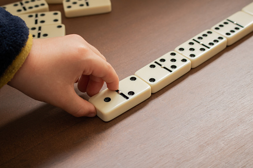 Child playing dominoes on wooden table. Detail of child's hand and domino tiles seen from above. Educational games, development and indoor activities concepts