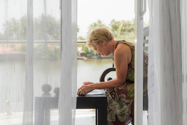 matura donna di 55 anni che lavora su un balcone all'aperto in un hotel contemporaneo. la vista da una stanza attraverso la porta aperta del balcone. - women mature adult 50 55 years 50s foto e immagini stock