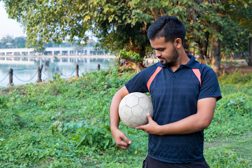 Outdoor Portrait of teenage soccer player holding a football in his hand and looking at camera. Young man during training on football field.