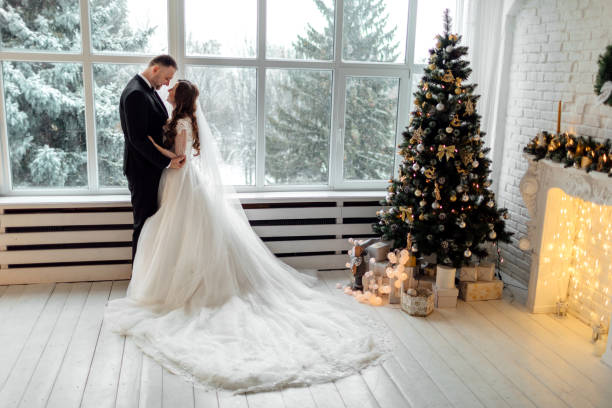 junges paar in liebe braut und bräutigam posiert im studio auf hintergrund mit weihnachtsbaum geschmückt in ihrem hochzeitstag zu weihnachten in der nähe des großen panoramafensters. - christmas wedding bride winter stock-fotos und bilder