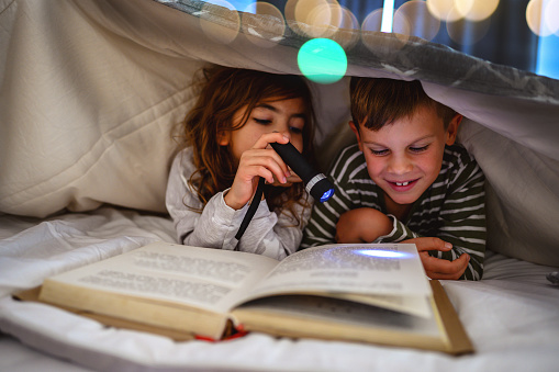 Two children are reading a book in bed under a sheet.They are hiding under a homemade playing tent, made of bedding. Little girl is reading a story book with a flashlight in her hand.