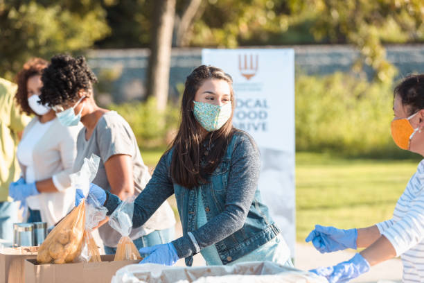 Young woman volunteers at outdoor food bank A young woman wears a protective face mask while volunteering at an outdoor food bank during the COVID-19 pandemic. She is sorting through food donations. charitable foundation photos stock pictures, royalty-free photos & images