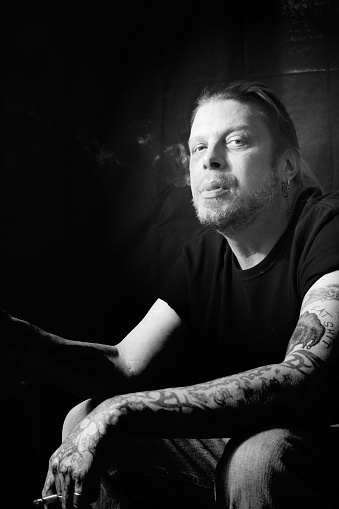 Studio shot of a man with tattoos smoking a cigarette looking at the light on a black background.