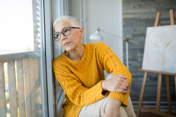 Mature artist sitting thoughtfully and looking through the window Portrait of a sad mature woman sitting on the floor of her living room and looking through the window. grey hair on floor stock pictures, royalty-free photos & images