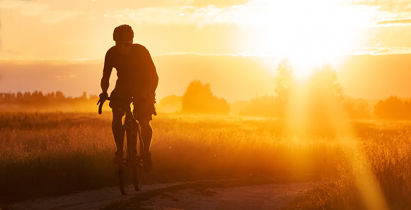 Silhouette of a cyclist on a gravel bike riding a trail in a field on a dramatic sunset background.
