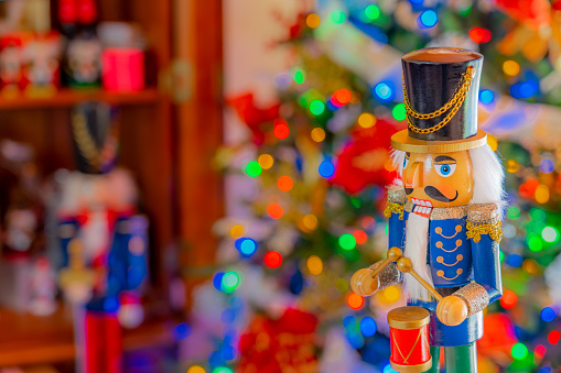 A Christmas nutcracker solider  beating a drum, stands in front of  a soft focus Christmas tree and cabinet.