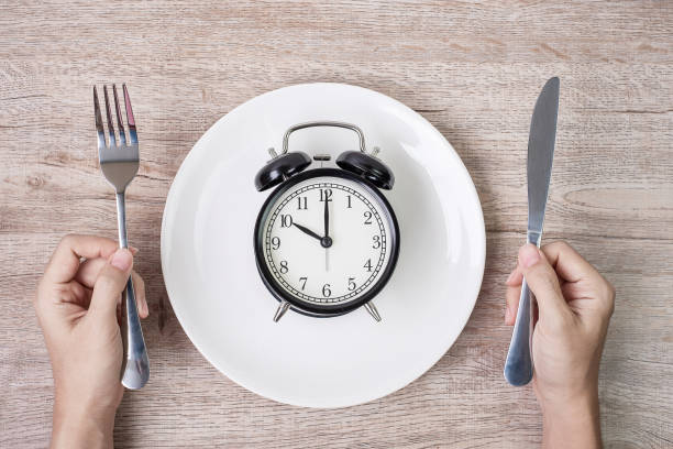 istock Hands holding knife and fork above alarm clock on white plate on wooden table background. Intermittent fasting, Ketogenic dieting, weight loss, meal plan and healthy food concept 1283129490