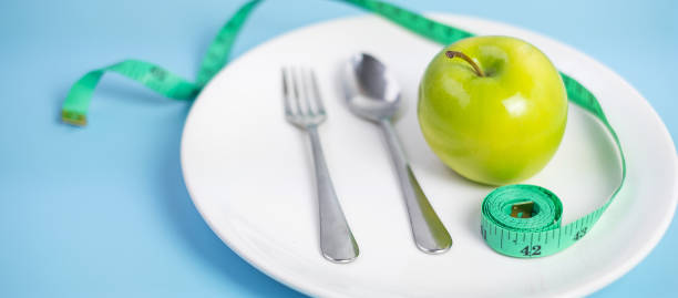 istock Top view spoon and fork, green apple on white ceramic plate with green Measuring tape on blue  background. dieting, weight loss, obesity and food control concept 1283129494
