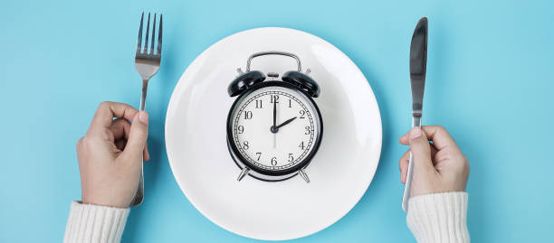 istock Hands holding knife and fork above alarm clock on white plate on blue background. Intermittent fasting, Ketogenic dieting, weight loss, meal plan and healthy food concept 1283129549