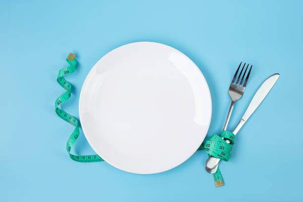 istock Top view Green Measuring tape wrapped around fork and knife with white ceramic dish on blue background. dieting, weight loss, obesity and food control concept 1283129614