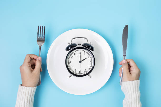 hands holding knife and fork above alarm clock on white plate on blue background intermittent