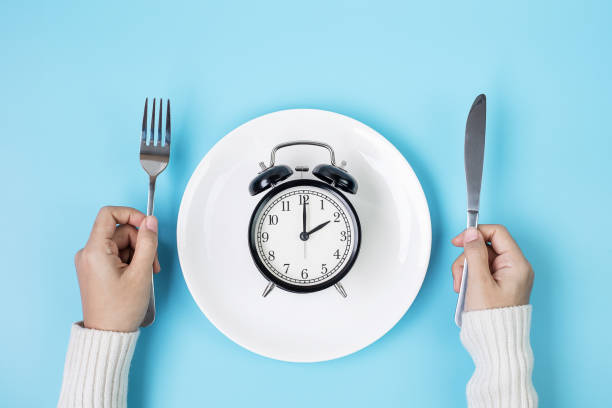 The Top Ten Health Benefits of Fasting: From Weight Loss to Longevity 1 hands holding knife and fork above alarm clock on white plate on blue background intermittent