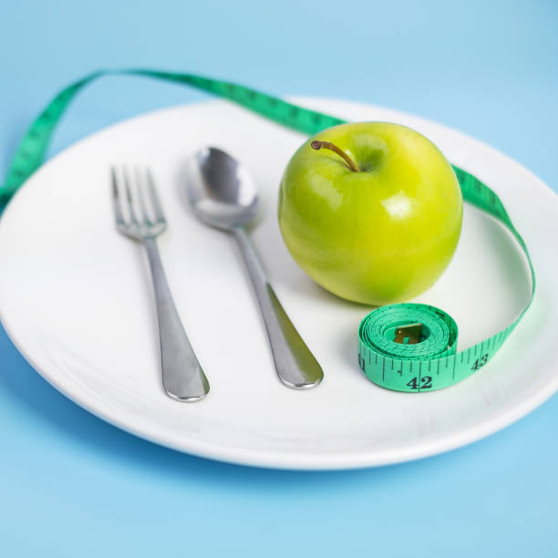 colher e garfo de vista superior, maçã verde na placa de cerâmica branca com fita de medição verde no fundo azul. dieta, perda de peso, obesidade e conceito de controle alimentar - plate blue loss tape measure - fotografias e filmes do acervo