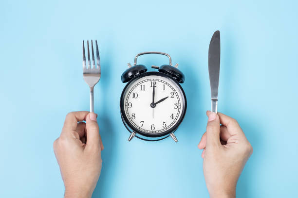 青い背景に白いプレートに目覚まし時計の上にナイフとフォークを持っている手。断続的な断食、ケト原性ダイエット、減量、食事プラン、健康的な食品コンセプト - plate blue dishware white ストックフォトと画像