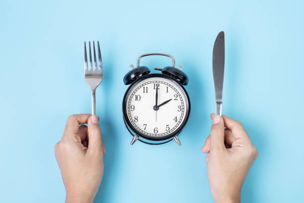 istock Hands holding knife and fork above alarm clock on white plate on blue background. Intermittent fasting, Ketogenic dieting, weight loss, meal plan and healthy food concept 1283129988