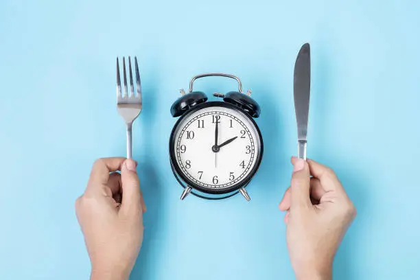 Hands holding knife and fork above alarm clock on white plate on blue background. Intermittent fasting, Ketogenic dieting, weight loss, meal plan and healthy food concept