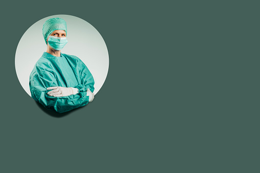 Mature female surgeon wearing mask and coveralls during COVID-19 pandemic. Portrait of confident doctor is with arms crossed. She is against green background.