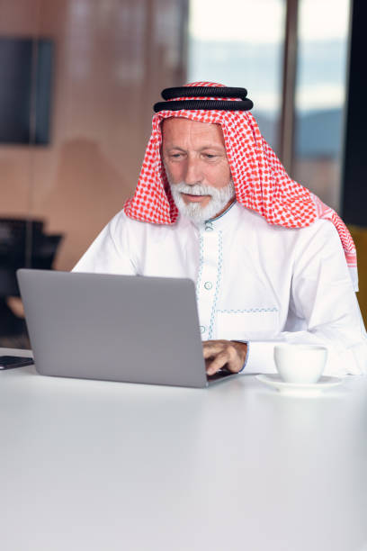 hombre de negocios árabe confiado y sonriendo en la oficina usando portátil. - headscarf islam senior adult east fotografías e imágenes de stock
