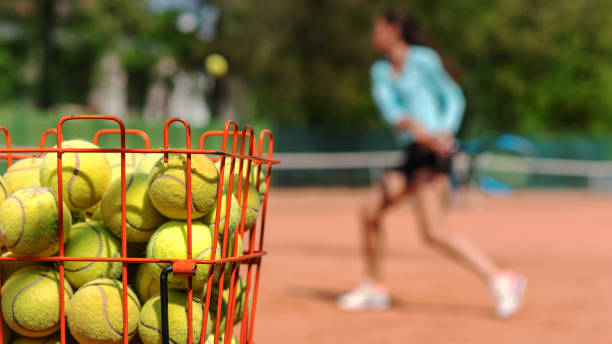 training process on game of tennis on clay court training process on game of tennis on clay courts in tennis academy, young athlete strikes backhand with racket on ball, foreground basket with tennis balls tennis ball container stock pictures, royalty-free photos & images