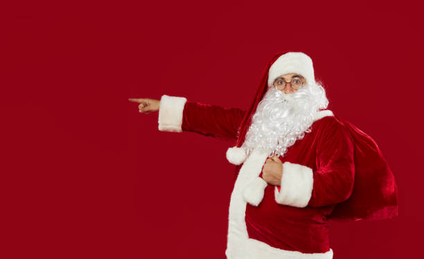 istock Surprised Santa Claus points to the left side for text. Red studio background 1283148797