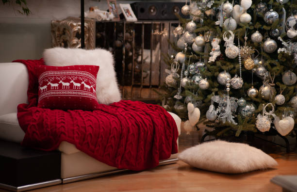 istock Christmas still life with red knit blanket and deers pattern cushion on a couch, white fluffy flooring pillow on a floor and decorated Christmas tree on the back 1283149509