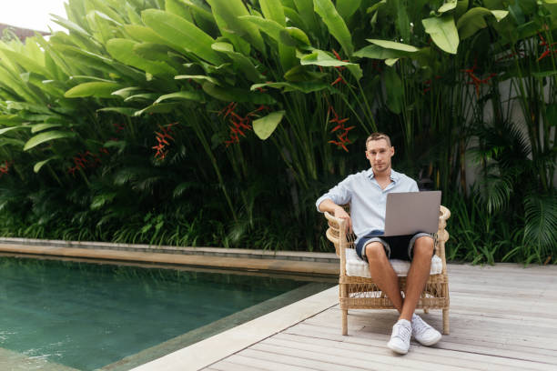 istock Young man using a laptop computer in a garden with a swimming pool. 1283149940