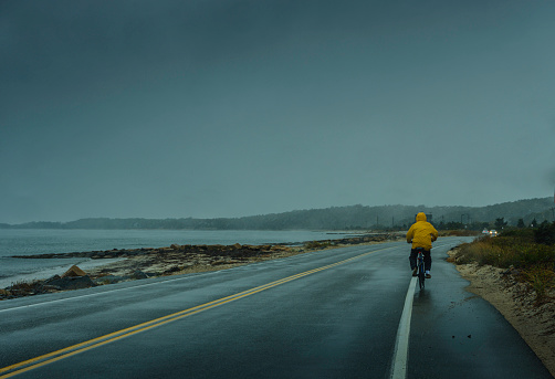 Biker cycling on the beach road in rainy weather. Passenger's view from a moving vehicle.