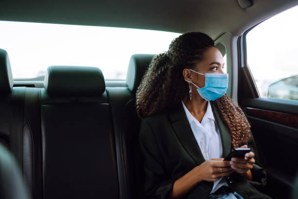 휴대 전화와 뒷좌석에 택시 차에 보호 의료 마스크를 편안한 여성 승객. - taint 뉴스 사진 이미지