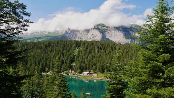 Photo of Turquoise colored lake Caumasee surrounded by forest reflected in the water near Flims, Grisons, Switzerland with funicular, restaurant and mountains in background.