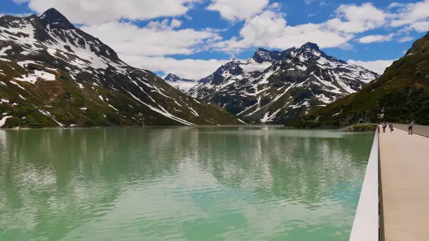 Photo of Tourists enjoying the beautiful view from Silvretta reservoir dam, Montafon, Austria with snow-capped mountains (including Biz Buin) in the background on partly cloudy day.