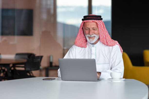 hombre de negocios árabe confiado y sonriendo en la oficina usando portátil. - headscarf islam senior adult east fotografías e imágenes de stock