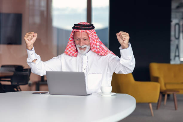 empresario árabe celebrando la victoria en la oficina usando portátil. - headscarf islam senior adult east fotografías e imágenes de stock