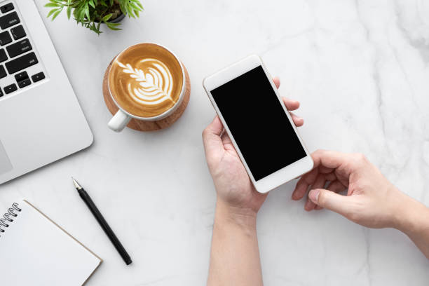 istock Hands holding white smartphone with blank screen over white marble office desk table with laptop computer, cup of coffee and office supplies. Top view, flat lay. 1283183571