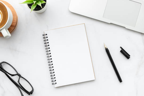 istock Blank notebook with pen are on top of white marble office desk table with laptop computer, cup of coffee and supplies. Top view, flat lay. 1283183673