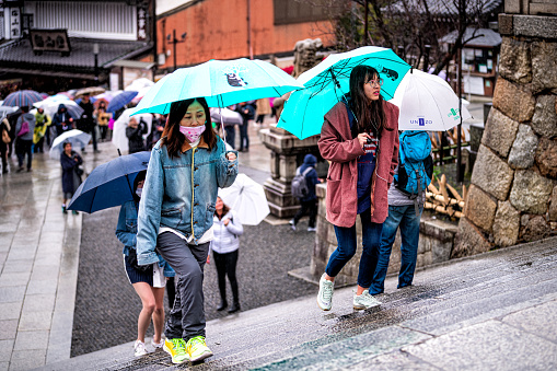 Kyoto, Japan - April 10, 2019: Many people with umbrellas walking up steps stairs in mask during rainy day by street near Kiyomizu-dera temple