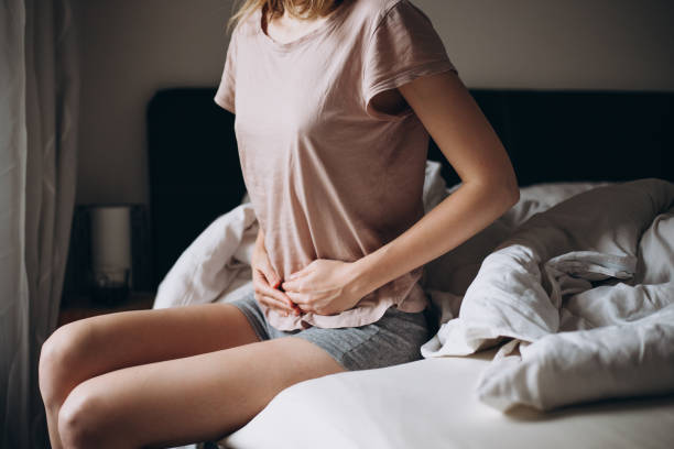 giovane donna che soffre di forte dolore addominale, tiro al coperto in stile casual. donna seduta a letto la mattina - menstruation foto e immagini stock