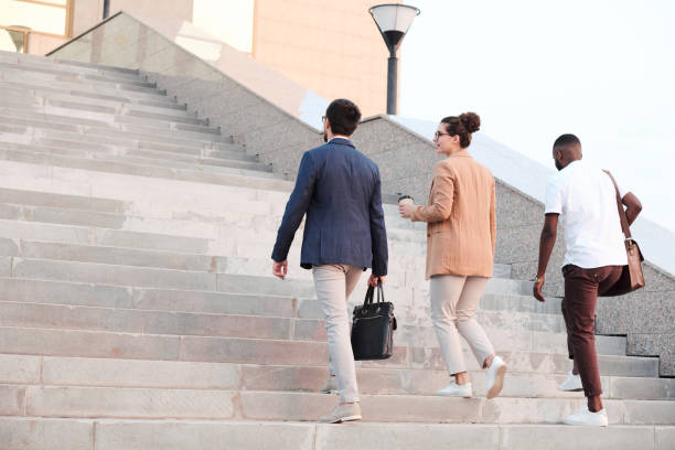 Rear view of business partners discussing new contract while moving upstairs Rear view of three young business partners in smart casualwear discussing terms of new contract while moving upstairs along riverside after office outdoor staircase stock pictures, royalty-free photos & images