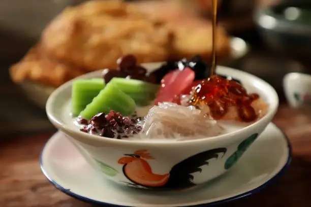 Che Hun Tiau, an icy dessert of Tapioca Cendol (noodle jelly) from Pontianak, West Kalimantan. The cendol is accompanied with Ketan Hitam (black glutinous rice), Bongko (pandan mung bean cake), Kacang Merah (red bean), Kolang Kaling (palm fruit) and Cincao (grass jelly) in coconut milk soup sweetened with palm sugar syrup. The syrup is being poured into the dish. This dessert dish is plated in a ceramic bowl with an underliner plate.