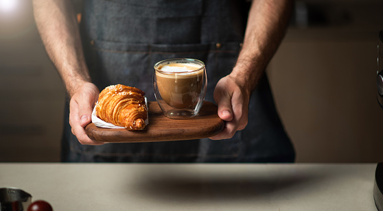 Male barista serving cup of coffee with croissant in a bar close up