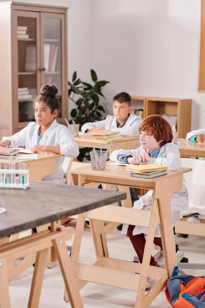 istock Group of elementary schoolkids in whitecoats sitting by desks in two rows 1283210917