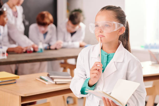 istock Pensive cute schoolgirl in whitecoat and protective eyeglasses holding copybook 1283210940